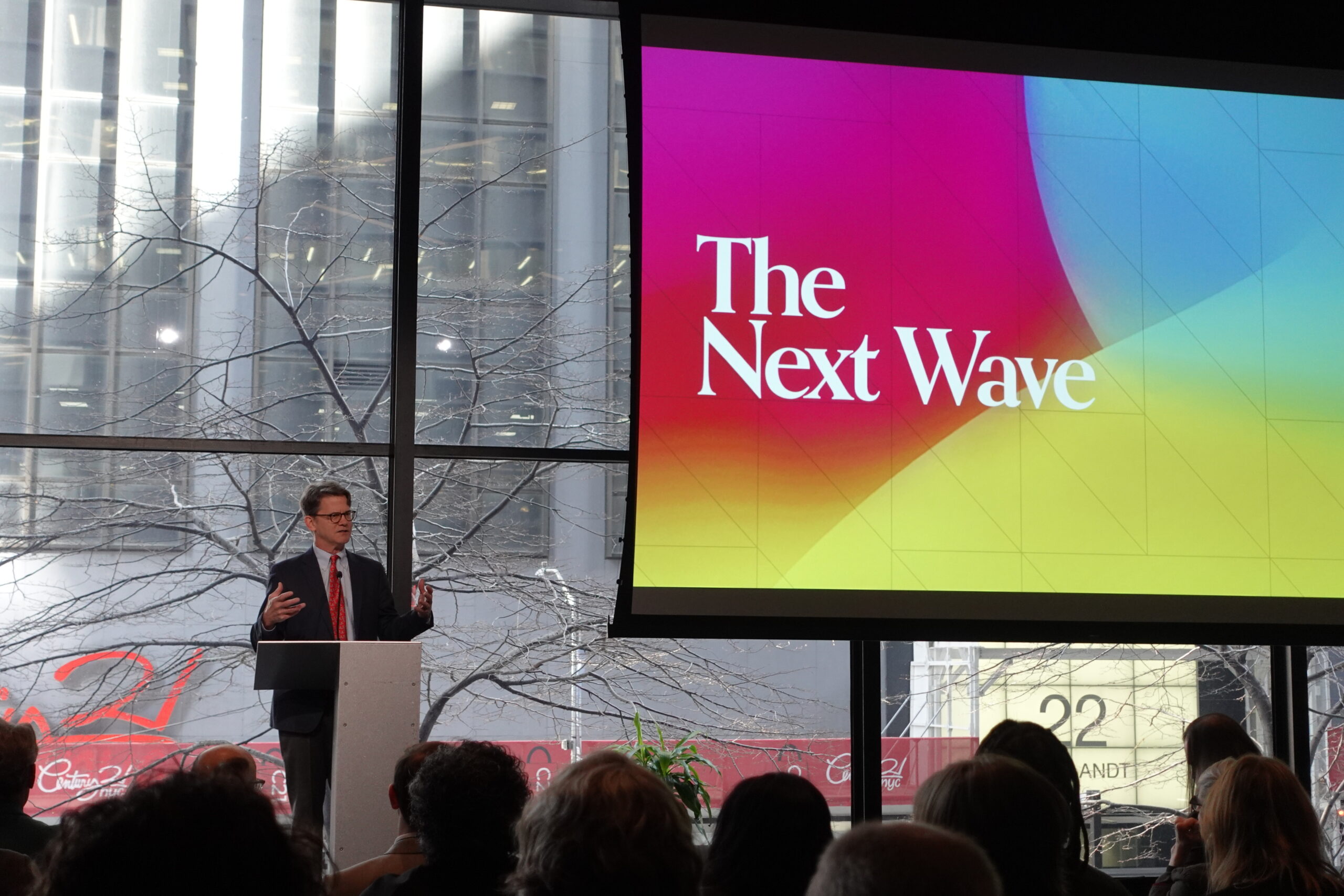 A speaker in a dark suit and red tie stands at a podium, addressing an audience at an indoor event. Behind him, a large screen displays the words "The Next Wave" in bold white text against a colorful gradient background. The event space features floor-to-ceiling windows showing an urban environment with leafless trees. The audience is seated in the foreground, facing the speaker.