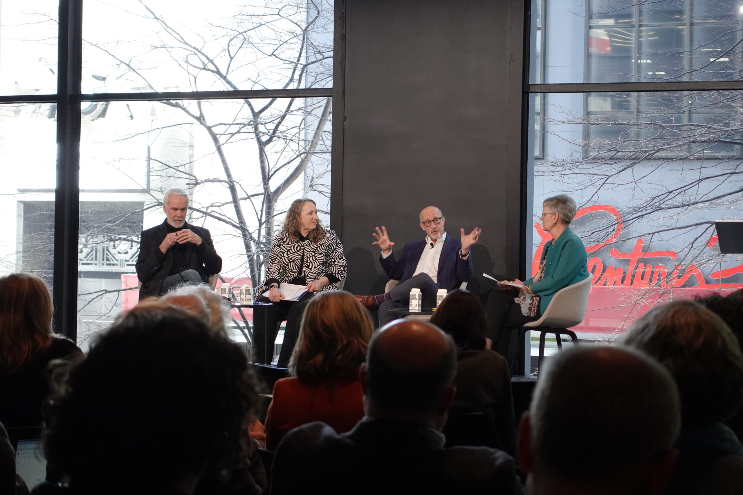 A panel discussion taking place in a modern event space with large windows overlooking an urban scene. Four panelists are seated on stage, engaged in conversation. One man in a navy suit gestures animatedly while speaking, while the others, including a woman in a teal blazer holding a notepad, listen attentively. The audience is visible in the foreground.