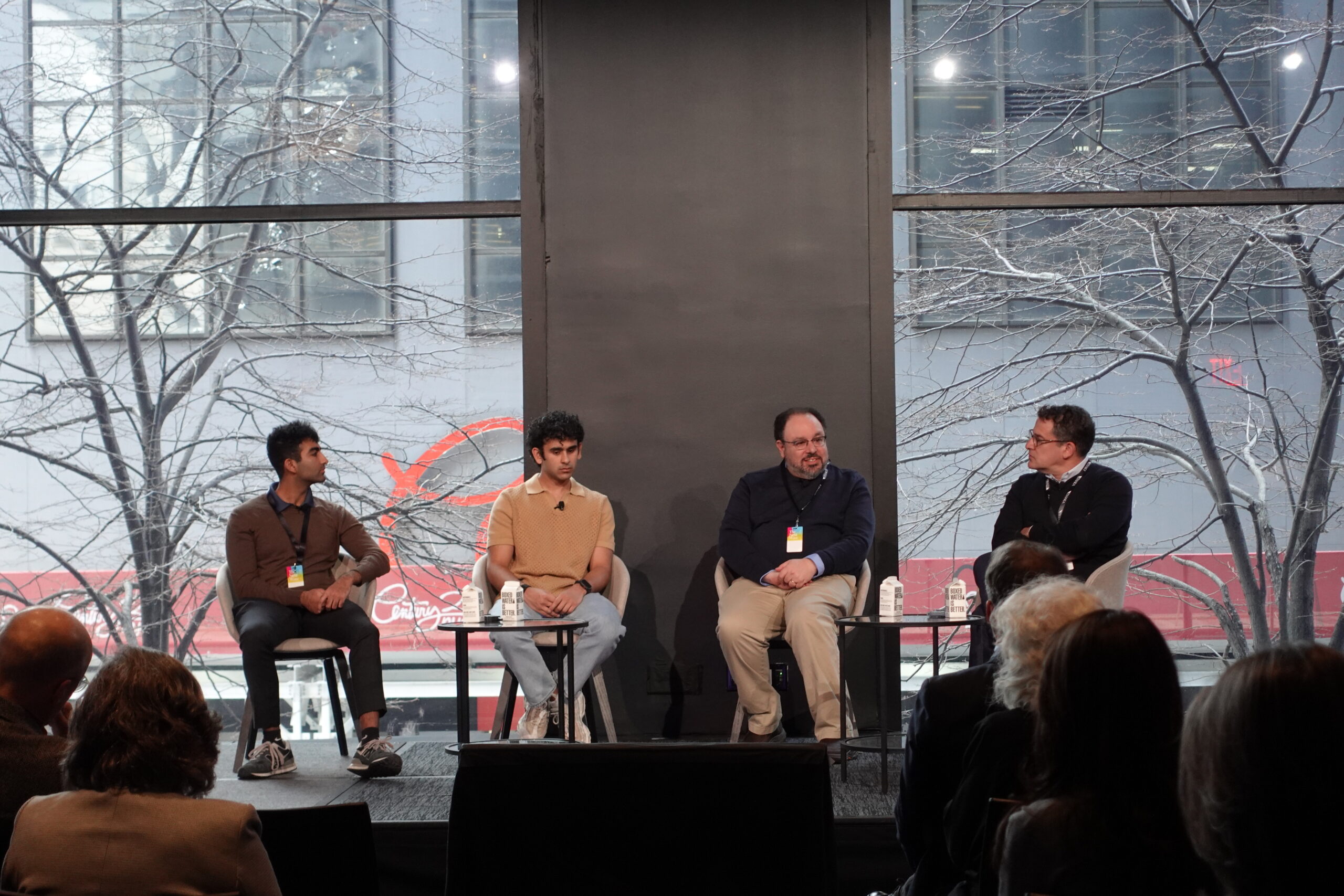 A panel discussion in a modern event space with large windows overlooking an urban scene. Four panelists are seated on stage, engaged in conversation. The audience is visible in the foreground.