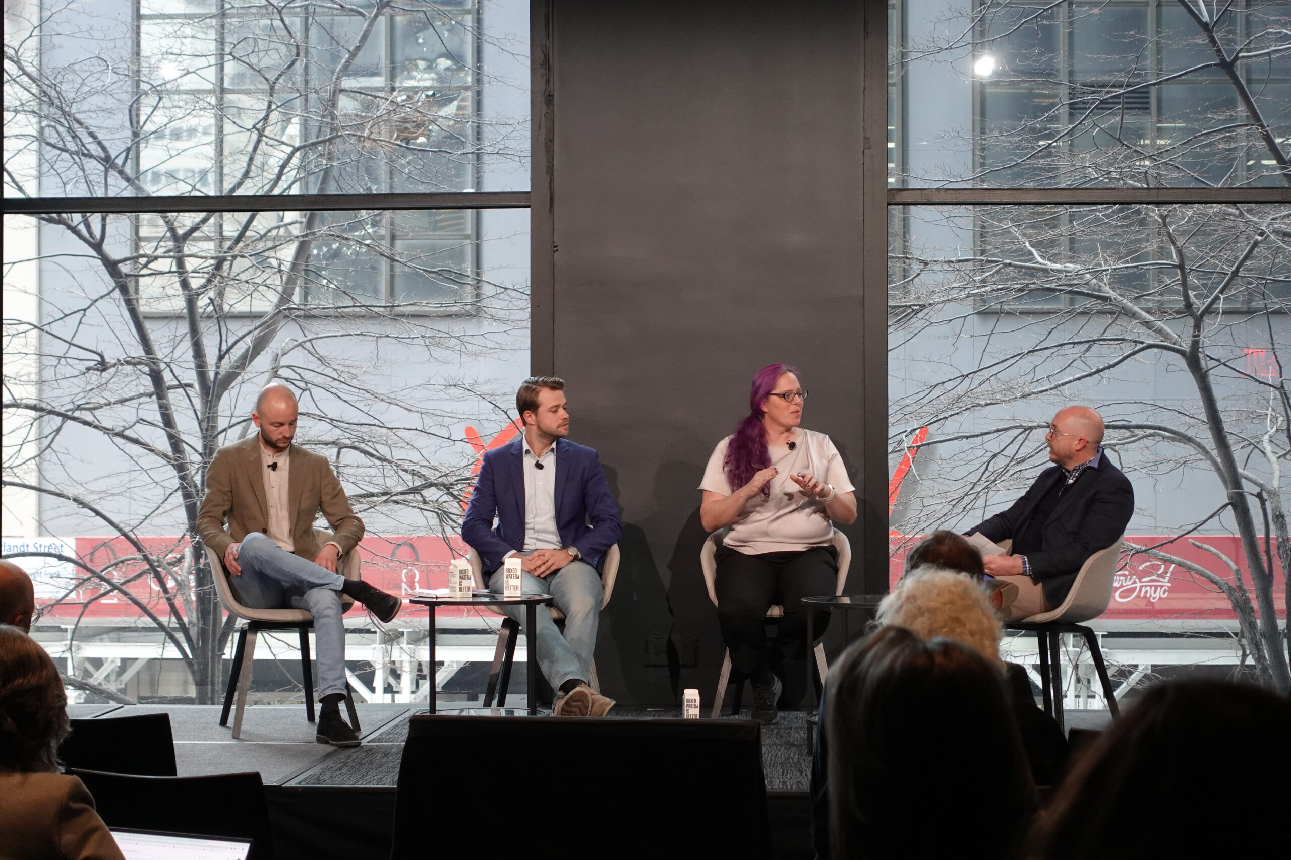 A panel discussion in a modern event space with large windows overlooking an urban scene. Four panelists are seated on stage, engaged in conversation. A woman with purple hair, wearing a light-colored shirt, is speaking and gesturing with her hands, while three male panelists in blazers listen. The audience is visible in the foreground.