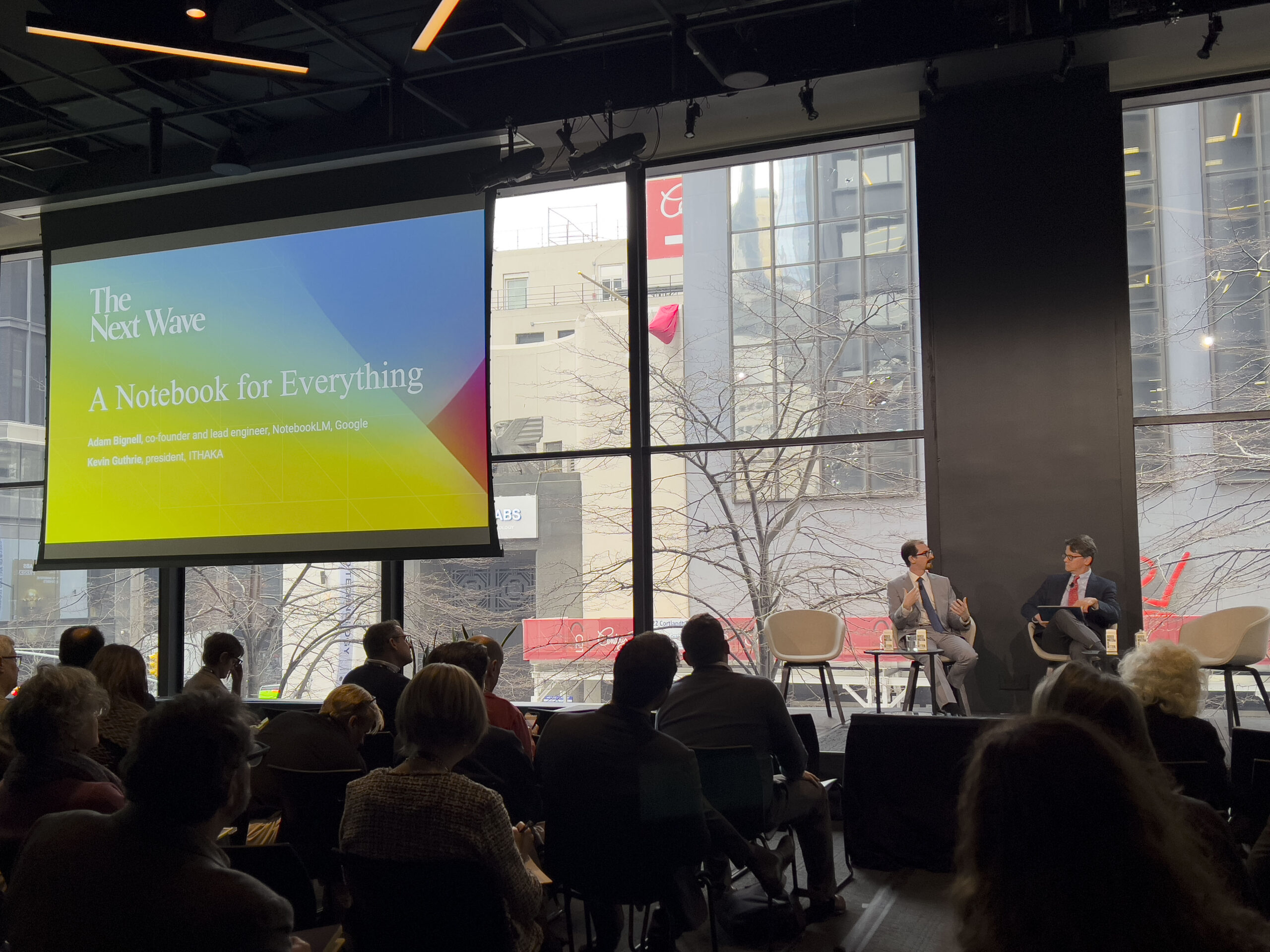 A panel discussion in a modern event space with large windows overlooking an urban cityscape. Two panelists in suits are seated on stage, engaged in conversation. A large screen above them displays a colorful slide titled The Next Wave: A Notebook for Everything, featuring the names Adam Bignell (Google) and Kevin Guthrie (ITHAKA). The audience is visible in the foreground, attentively watching the discussion.